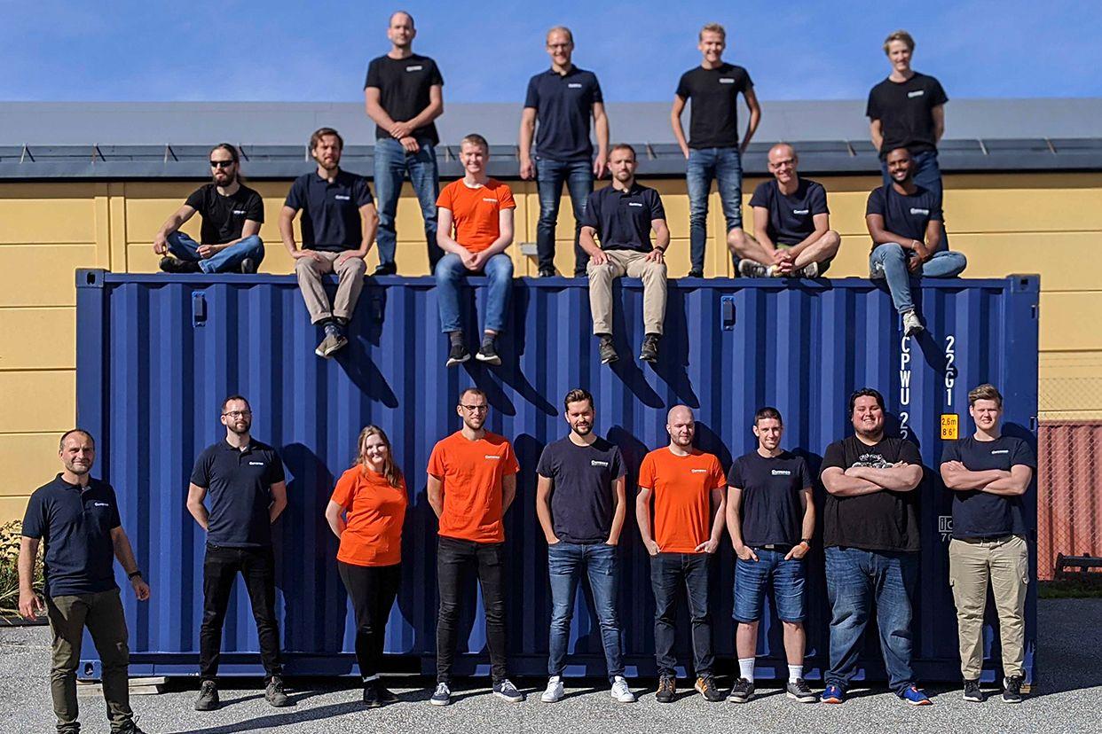 Group photo of 19 Solwr employees sitting on top, and standing in front of, a big blue storage container